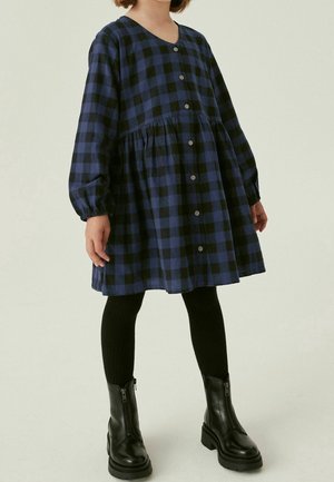 Robe à carreaux bleu marine et noir avec manches longues, boutonnée à l'avant, et taille à basque, associée à des collants côtelés noirs et des bottines noires.