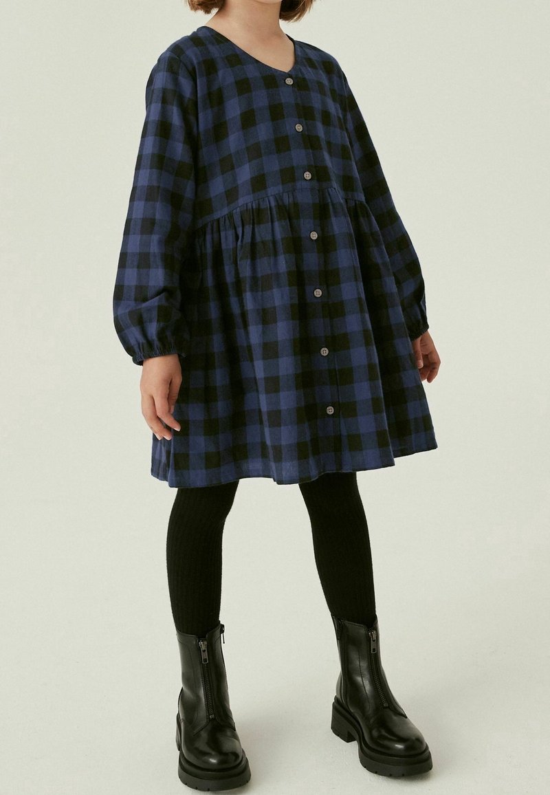 Robe à carreaux bleu marine et noir avec manches longues, boutonnée à l'avant, et taille à basque, associée à des collants côtelés noirs et des bottines noires.