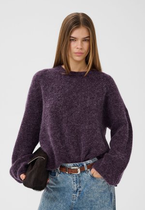 Jeune femme portant un pull violet duveteux et un jean bleu, tenant une pochette en fourrure foncée, une main dans la poche.