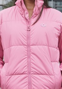 Rosa vadderad jacka med hög krage, dragkedjestängning och en broderad logotyp på bröstet. Slät yta, vadderad design.