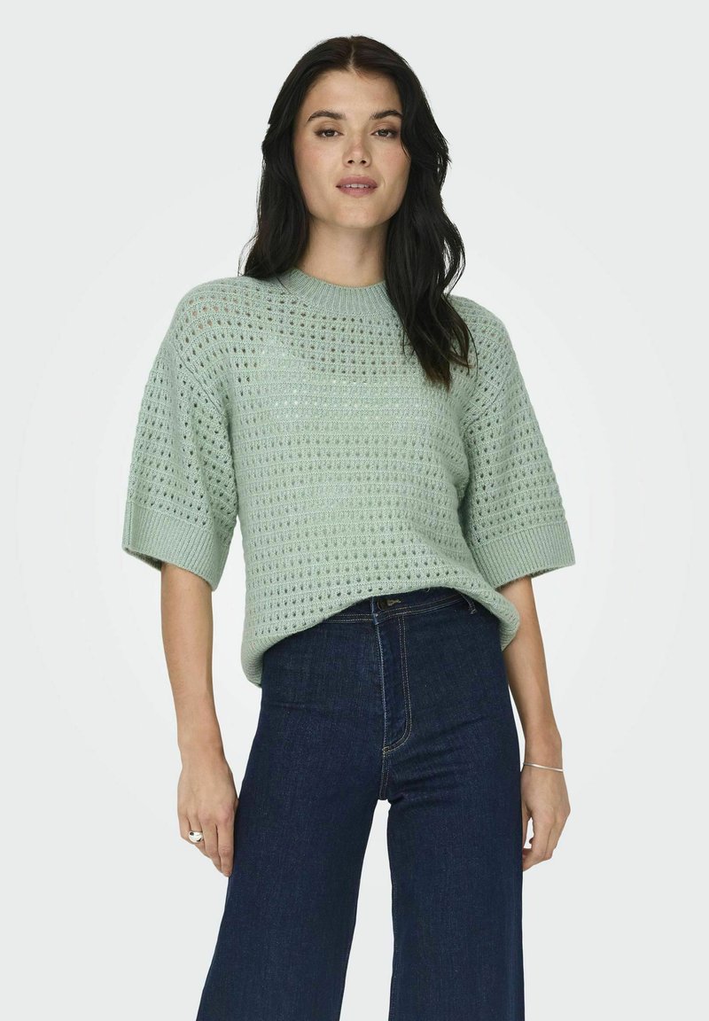 Maglione lavorato a maglia verde chiaro con una vestibilità ampia, caratterizzato da un motivo testurizzato e maniche corte, abbinato a jeans in denim scuri a gamba larga.