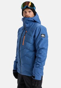 Mann in einer blauen Winterjacke mit Kapuze und orangefarbendem Reißverschluss, schwarzen Handschuhen, schwarzer Mütze und Skibrille auf der Stirn, stehend vor einem weißen Hintergrund.