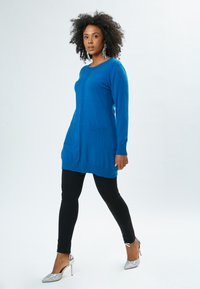 Cardigan blu in maglia con maniche lunghe, due tasche frontali e un motivo verticale testurizzato. Abbinato a leggings neri e tacchi argentati.