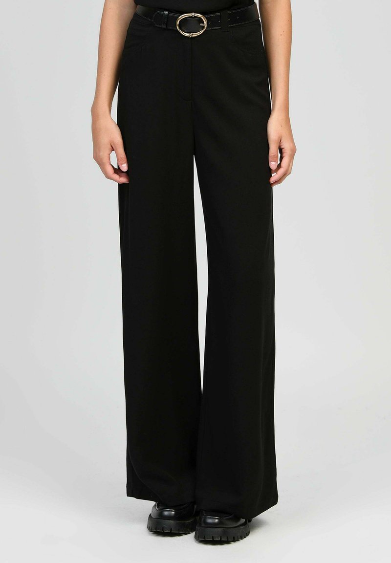 Pantalons larges noirs en tissu doux, avec une taille ajustée et une boucle rond en métal doré sur une ceinture noire. Design élégant et ajusté.