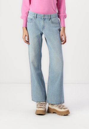 Femme portant un jean large bleu clair, un pull à manches longues rose et des bottes beige doublées de fourrure, debout devant un fond blanc.