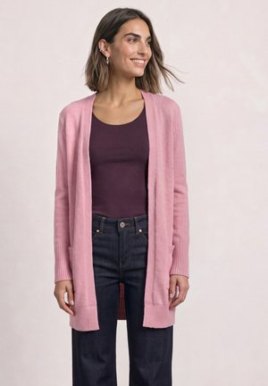 Femme souriante portant un cardigan rose ouvert devant, un haut violet foncé et un jean bleu foncé sur un fond clair uni.