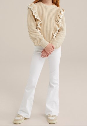 Fille portant un pull en maille beige avec des volants verticaux, un pantalon blanc évasé et des baskets beiges, debout les mains jointes.