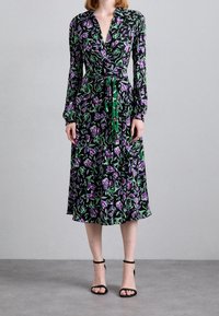 Černé zavinovací šaty s květinovým vzorem v purpurových a zelených akcentů, dlouhými rukávy a staženým pasem svázaným zeleným páskem.