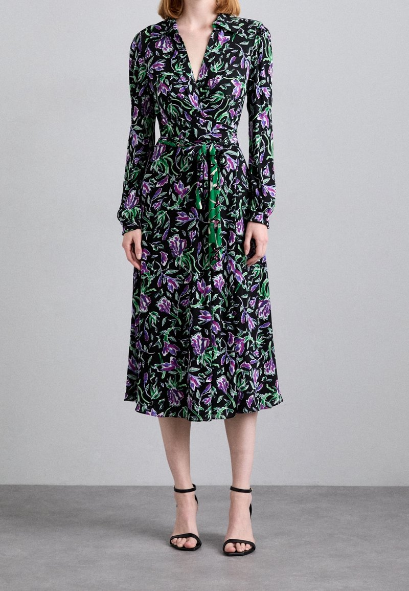 Černé zavinovací šaty s květinovým vzorem v purpurových a zelených akcentů, dlouhými rukávy a staženým pasem svázaným zeleným páskem.