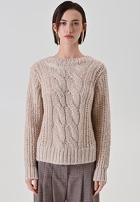 Maglione beige lavorato a maglia con motivo a treccia, orlo e polsini a costine e texture. Tessuto morbido e spesso, collo tondo. Indossato sopra pantaloni a quadri.
