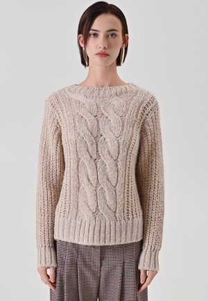 Maglione beige lavorato a maglia con motivo a treccia, orlo e polsini a costine e texture. Tessuto morbido e spesso, collo tondo. Indossato sopra pantaloni a quadri.