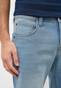 Jasnoniebieskie dżinsy mają klasyczny pięciokieszeniowy fason z widocznym przeszyciem i lekko wyczuwalnym efektem wyblaknięcia. Widać dopasowany krój.