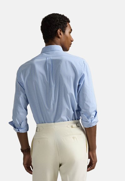 Chemise rayée bleu clair avec col à boutons et manches retroussées, assortie à un pantalon crème avec poches arrière et ajusteurs latéraux.