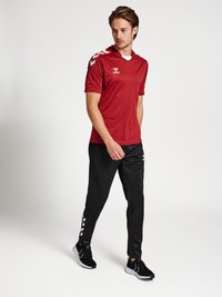 Fiatal férfi piros sportmezt és fekete, fehér cikk-cakk mintás melegítőnadrágot visel, fehér háttér előtt sétál.