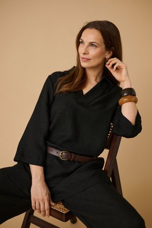 Vrouw met lang bruin haar, gekleed in een zwarte outfit en een bruine riem, zit op een houten stoel, rust haar hand op haar gezicht en kijkt opzij.