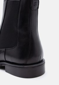 Μαύρο δερμάτινο μποτάκι Chelsea με ελαστική πλαϊνή επένδυση και ραμμένες λεπτομέρειες σε λευκό φόντο.