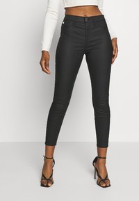 Vaqueros skinny negros de cintura alta, fabricados con material elástico, que presentan un cierre frontal con botón, un diseño elegante y un ajuste hasta el tobillo, combinados con tacones negros de tiras.