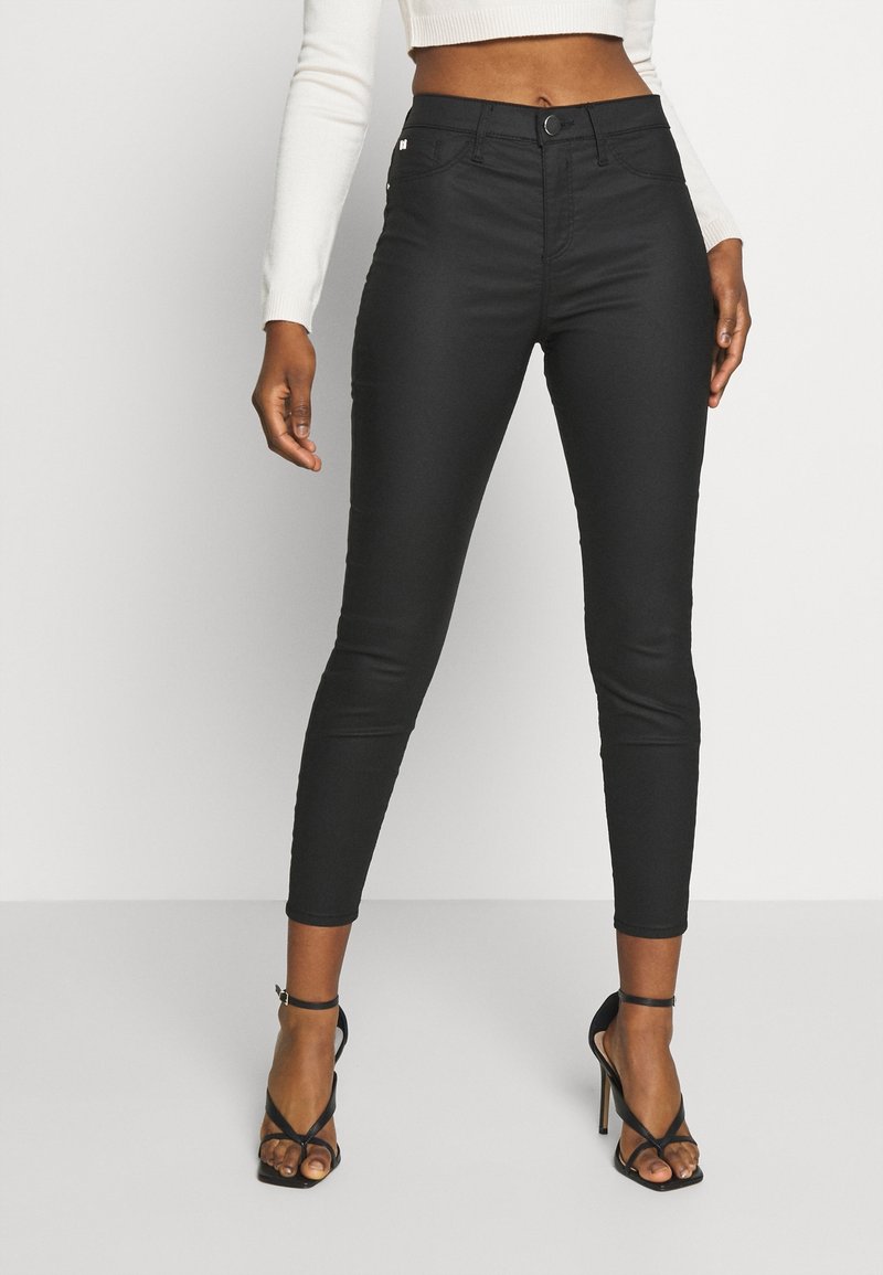 Vaqueros skinny negros de cintura alta, fabricados con material elástico, que presentan un cierre frontal con botón, un diseño elegante y un ajuste hasta el tobillo, combinados con tacones negros de tiras.