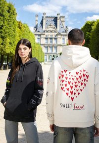 Deux personnes à l'extérieur devant un bâtiment historique ; l'une porte un sweat à capuche noir avec des motifs, l'autre un sweat à capuche blanc avec des motifs de cœurs rouges et « SWEET heart ».