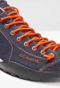 Farkkukenkä, jossa on oranssit nauhat, harmaat reiät ja musta kumipohja. Sivussa pronssinvärinen SCARPA-logo.