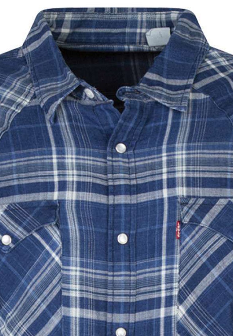 Chemise en flanelle à carreaux bleus avec des boutons argentés, col pointu, et deux poches avant arborant une étiquette de marque sur la poche gauche. Tissu texturé.