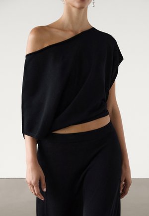 Woman wearing a loose black off-shoulder top and matching black pants with dangling earrings against a plain background.