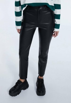 Person trägt hoch taillierte schwarze Kunstlederhose, gestreiftes langärmeliges Shirt und klobige schwarze Plateausneaker, steht vor einem schlichten Hintergrund.