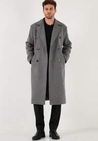 Uomo in piedi con le mani in tasca, indossa un lungo cappotto grigio doppiopetto, pantaloni neri, camicia nera e scarpe nere su sfondo bianco.