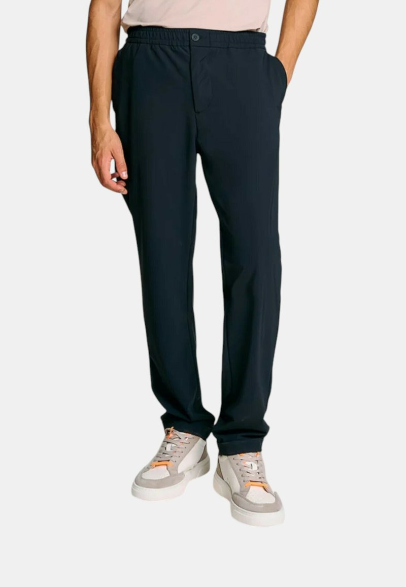 Uomo che indossa pantaloni casual neri con vita elastica e sneaker bianche con dettagli arancioni, in piedi con una mano in tasca.