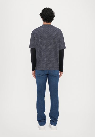 Īsa piedurkņu rakstota krekla zilā krāsā ar ģeometrisku rakstu, apvienots ar ziliem džinsiem un baltām kedām. Muguras skats parāda krekla brīvo piegriezumu.