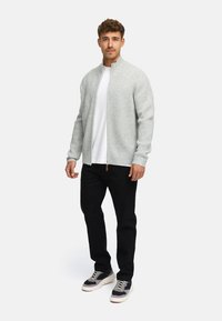 Suéter de punto gris claro con cremallera y textura acanalada, combinado con una camiseta blanca, jeans negros y zapatillas grises con acentos multicolores.