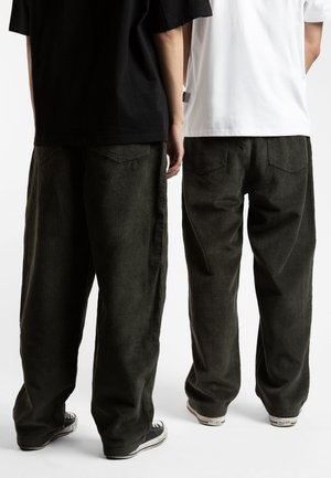 To personer iført sorte kordfløjlsbukser og sneakers står ved siden af hinanden, den ene i en sort trøje og den anden i en hvid trøje, med ryggen til.