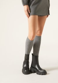 Botas negras de tobillo con suelas gruesas, de material de cuero suave, combinadas con calcetas gris jaspeadas hasta la rodilla. Diseño simple y acentos de hardware resistente.