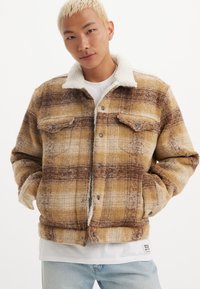Chaqueta a cuadros en tonos marrón y amarillo con cuello de sherpa, grandes bolsillos frontales, cierre de botones y un acabado de tela texturizada.