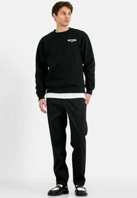 Zwart crewneck sweatshirt met opgedrukt logo, laag over een wit shirt, gecombineerd met zwarte broek en zwarte schoenen met witte accenten.