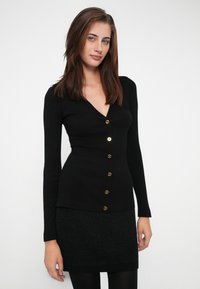 Black ribbed cardigan with a deep V-neck, gold buttons down the front, and long sleeves. Worn over a textured black skirt.