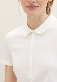 Vit polo t-shirt med klassisk krage, korta ärmar och en knappslå med sex matchande knappar. Texturerad tyg med ett subtilt mönster.