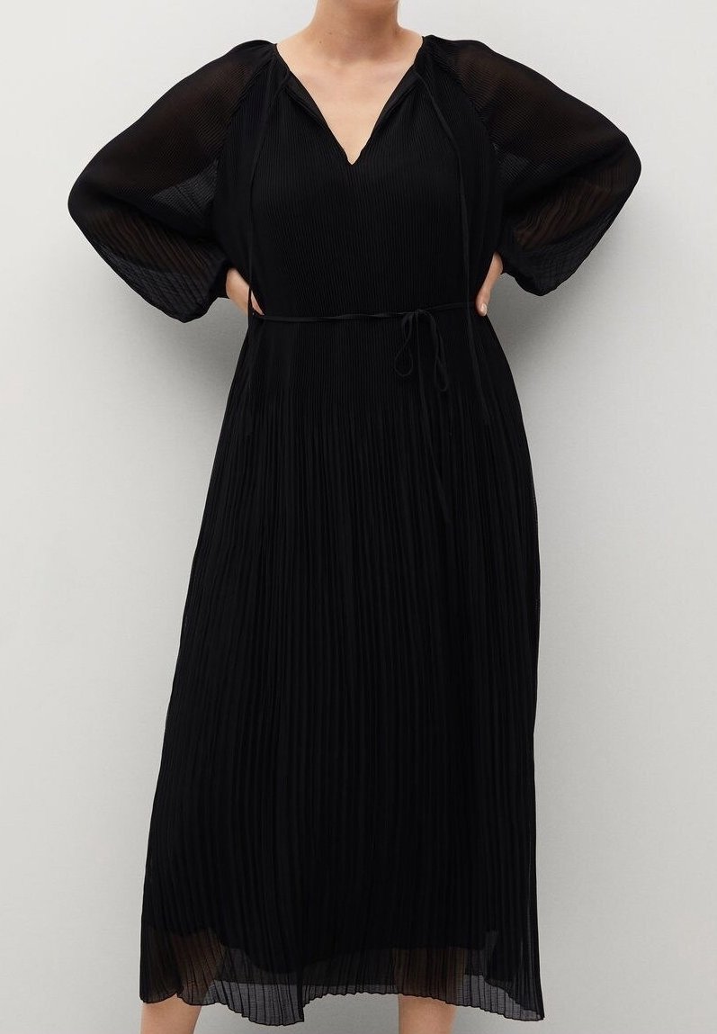 Femme portant une robe midi plissée noire avec des manches longues évêques transparentes et une fine ceinture à la taille, debout les mains sur les hanches.