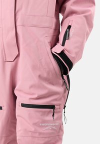 Salopette impermeabile rosa realizzata in tessuto resistente, con tasche a zip nere e il logo "Swedemount" in bianco sulla coscia anteriore.