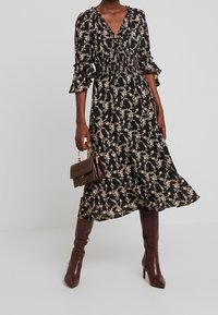 Black floral dress with a V-neck, smocked waist, and ruffled cuffs, paired with knee-high burgundy boots and a small brown handbag.