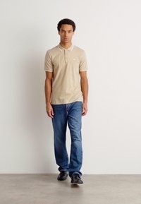 Beige polo skjorte med hvit krage og knapper, parret med blå jeans og svarte sko. Modell som står på en nøytral bakgrunn.