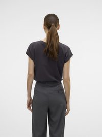 Femme aux cheveux bruns en queue de cheval, portant un haut gris foncé à manches courtes et un pantalon gris taille haute, se tenant devant un fond blanc uni.
