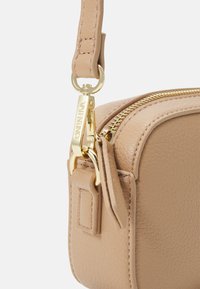 Kompakt beige lærveske med teksturert overflate, gullfarget hardware og glidelåslukking med avtagbar stropp.