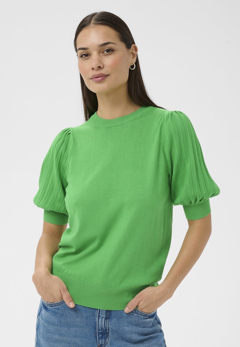 Femme aux longs cheveux foncés portant un pull vert vif à manches courtes bouffantes et un jean bleu, les mains dans les poches, fond neutre.