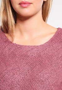 Maglione rosa con texture, caratterizzato da un ampio scollo e un sottile motivo a spina di pesce, realizzato in tessuto morbido e leggermente peloso.