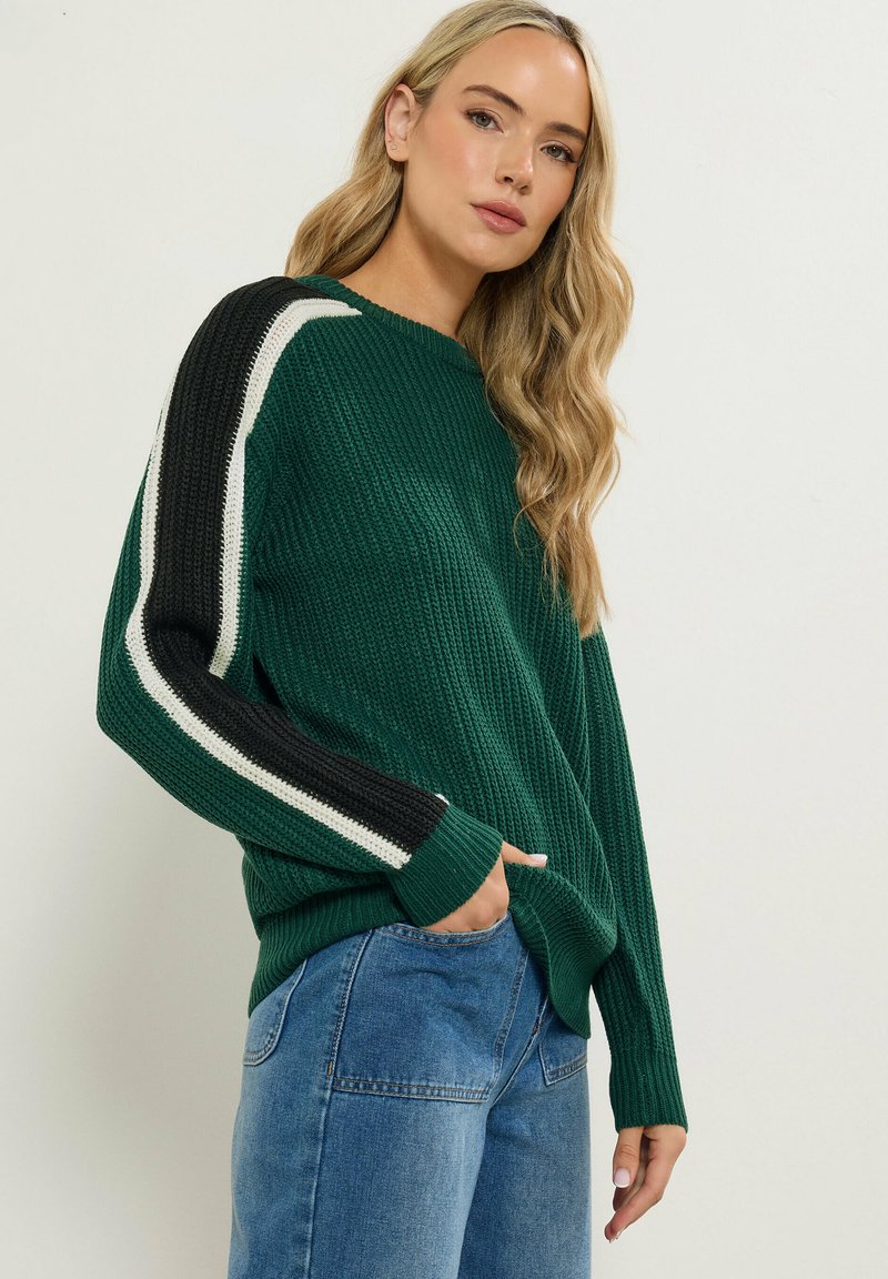 Maglione verde lavorato a maglia con maniche a strisce bianche e nere in contrasto, caratterizzato da una vestibilità rilassata e tasche laterali. Dettaglio del tessuto testurizzato visibile.