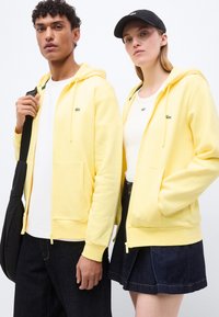 Deux personnes portant des sweats à capuche zippés jaunes assortis et des hauts blancs ; l'une tient une sangle de sac noire, l'autre porte une casquette noire et une jupe en jean.