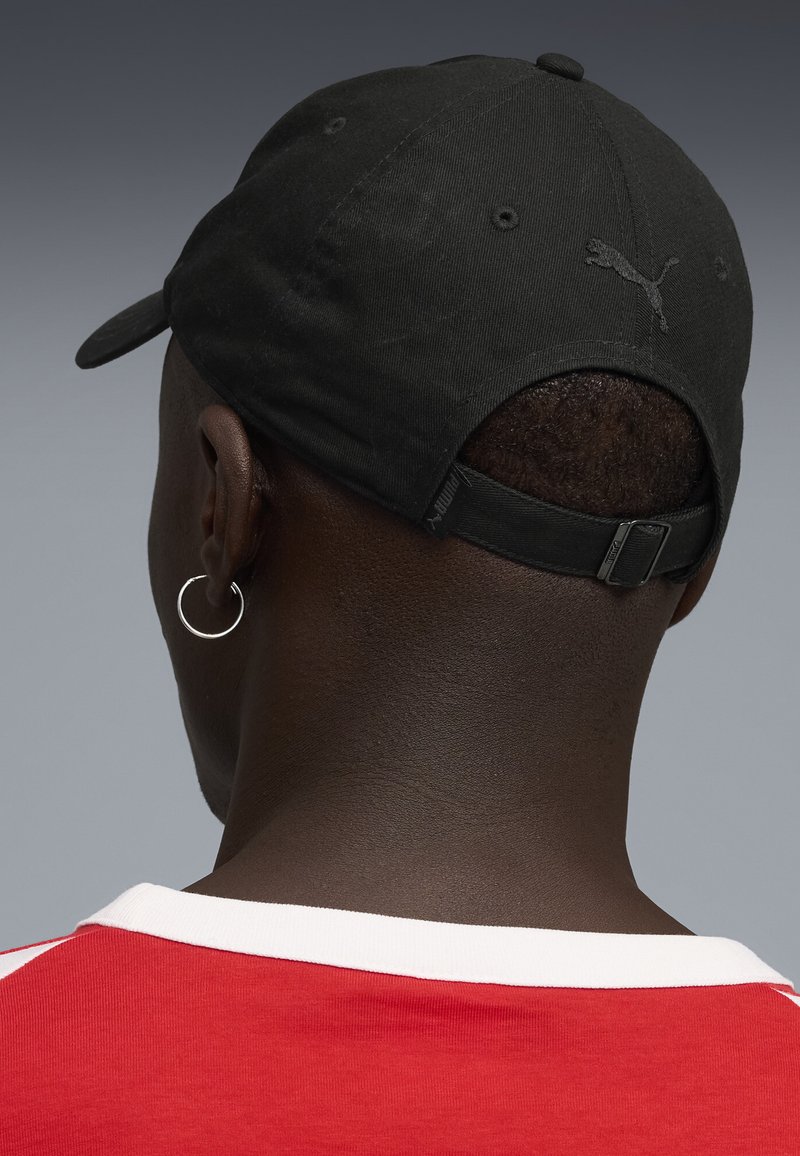 Homme portant une casquette noire Puma et une boucle d'oreille argentée en forme d'anneau, vu de dos, habillé d'une chemise rouge avec un col blanc sur fond gris.