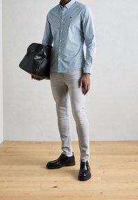 Světlomodrá kostkovaná košile na knoflíky, šedé úzké džíny, černé kožené boty a černá kožená taška, stojící na dřevěné podlaze.