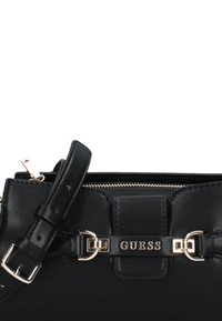 Borsa a tracolla in pelle nera con dettagli in metallo dorato, chiusura con zip e tracolla staccabile. Logo "GUESS" ben visibile.